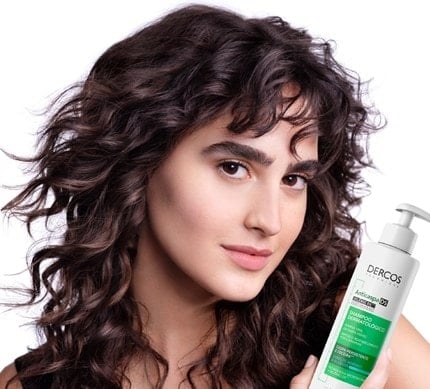 Modelo com cabelo brilhoso e ondulado, segurando um produto Dercos.
